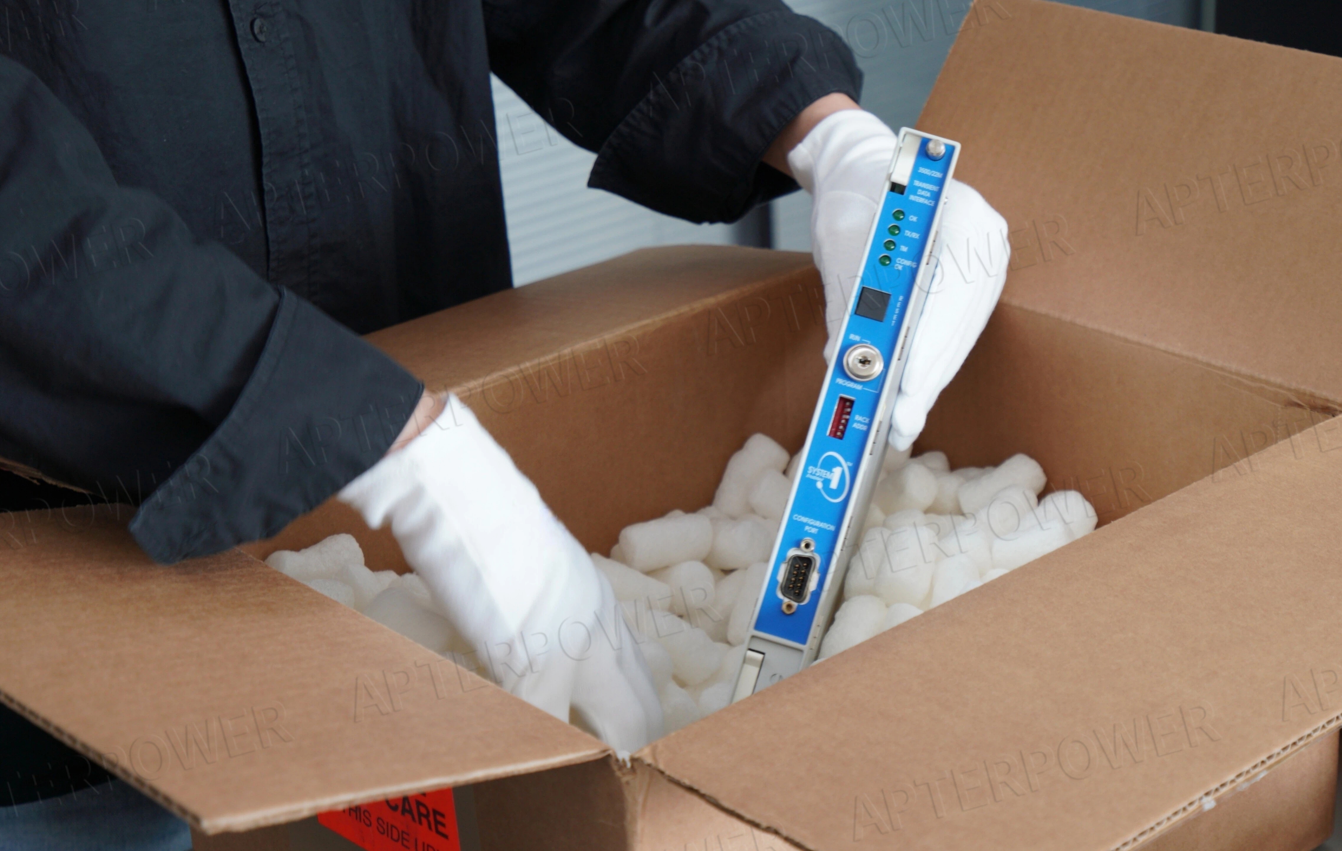 Gemini said A person wearing white gloves carefully placing a blue electronic module into a cardboard shipping box filled with packing peanuts.