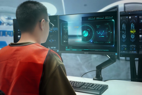 A technician in an orange safety vest intently monitoring complex data visualizations across multiple computer screens in a high-tech control room.